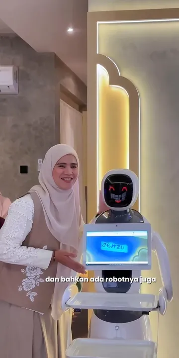 Nia Mahpud tersenyum di samping robot penyambut di Senzu Reflexology